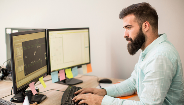 A man is wearing an aqua colour shirt with white checks. He is sitting at his desk and working on two computer monitors.