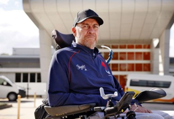 Daniel is wearing a blue jacket and black cap. He is looking at the camera. There is a building and two cars behind him