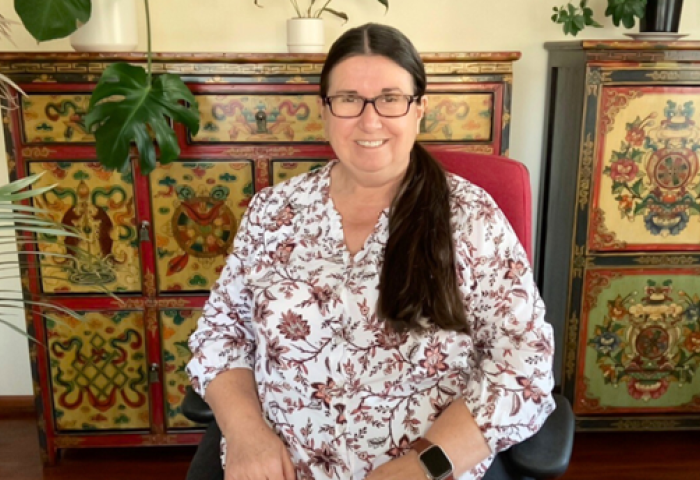 Sharon is smiling facing the camera, sitting in her red and black office chair. She is wearing a floral-patterned top.