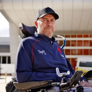 Daniel is wearing a blue jacket and black cap. He is looking at the camera. There is a building and two cars behind him
